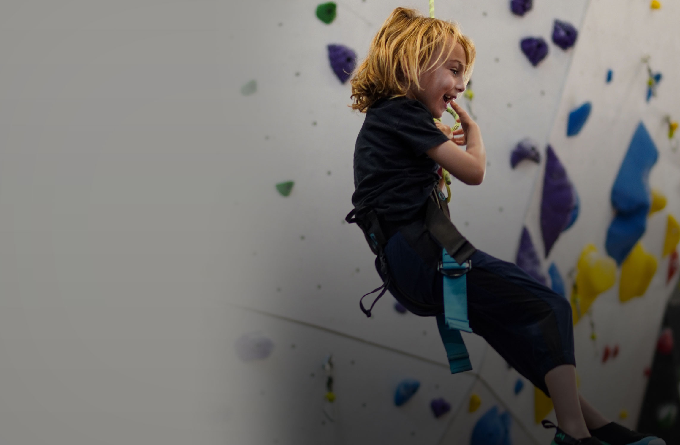 A smiling child with light brown hair, wearing a harness, sits suspended in an indoor rock climbing gym with colorful climbing holds visible in the background.