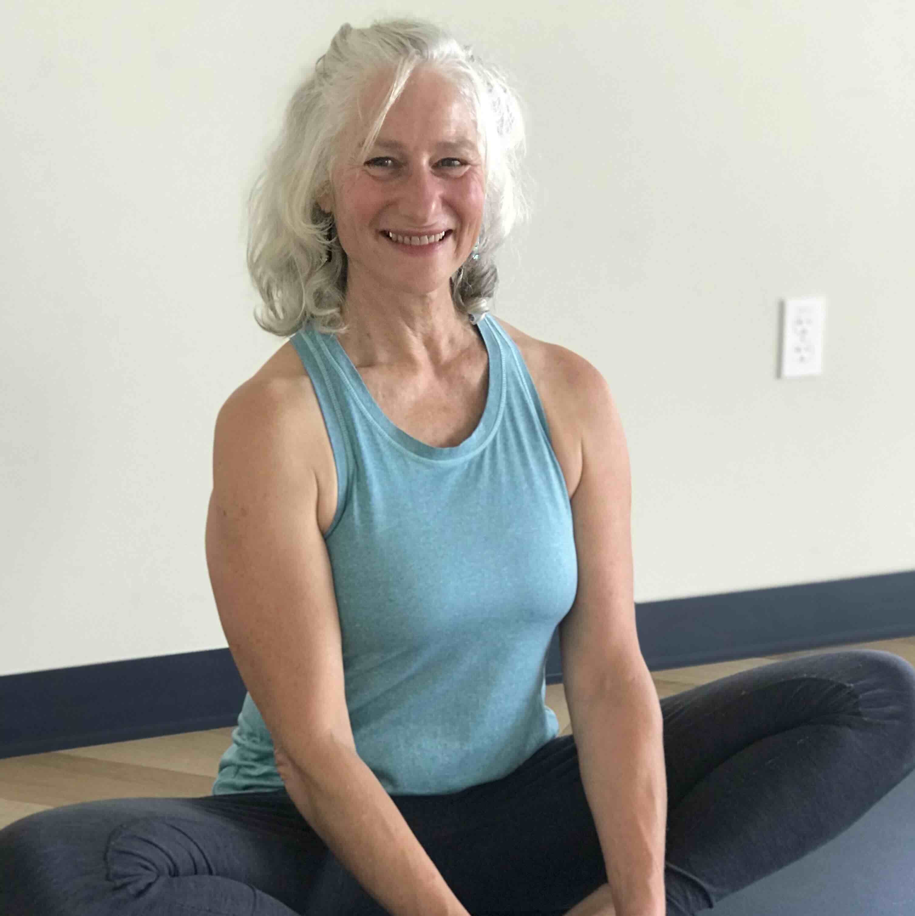 IMG_4805 A smiling older woman with white hair sits cross-legged on a yoga mat, wearing a blue sleeveless top and dark leggings, against a light-colored wall.
