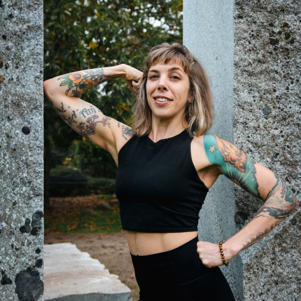 Lee square photo A woman with shoulder-length hair and colorful arm tattoos poses confidently outdoors, flexing her right arm and smiling. She wears a black sleeveless top and stands between two large stone pillars.
