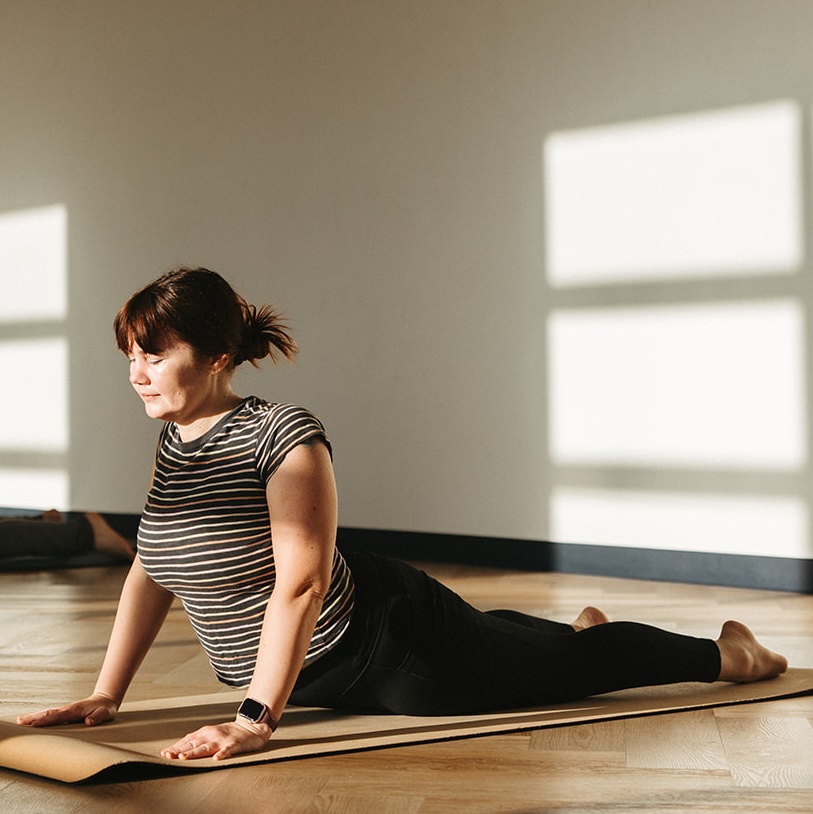 Session Woman on a yoga mat indoors, doing an upward-facing stretch with her arms straight and torso lifted. She wears a striped shirt, dark pants, and a smartwatch. Sunlight shines through windows onto the wooden floor.