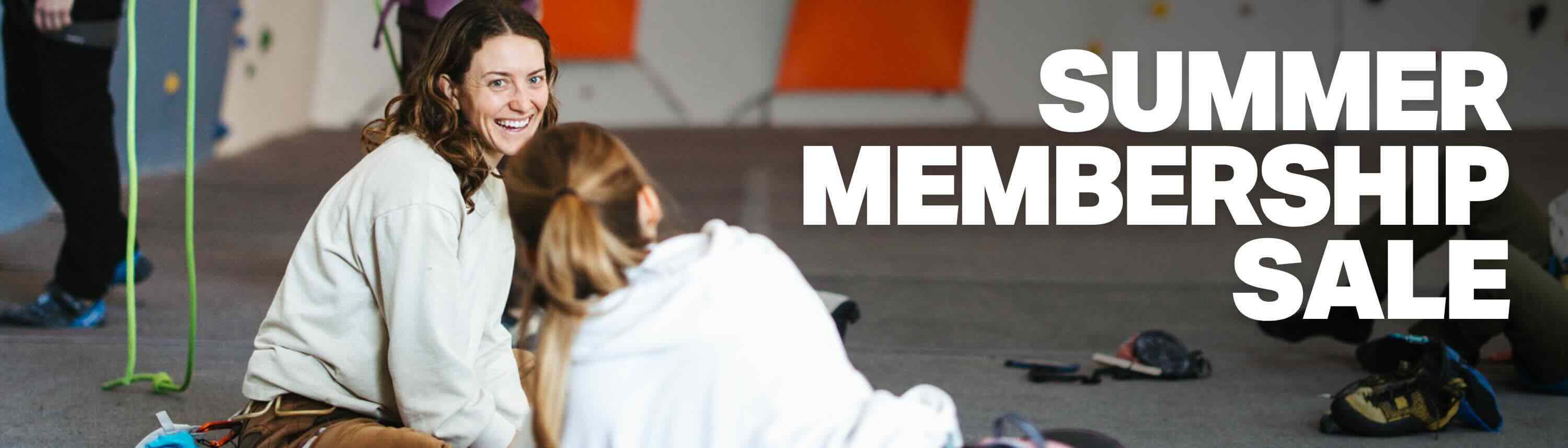 Two people sit on the floor of an indoor climbing gym, smiling and talking. Bold white text on the right reads: Summer Membership Sale. Climbing gear and ropes are visible in the background.