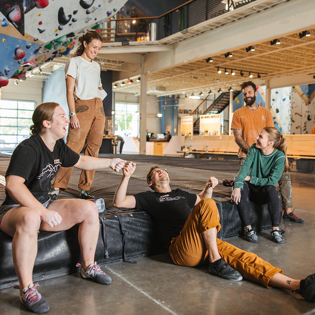 Session Five people are at an indoor rock climbing gym. Four are standing or sitting on mats and smiling, while one person lies on the mat, appearing playful or triumphant, as the group interacts cheerfully. Climbing walls are visible in the background.