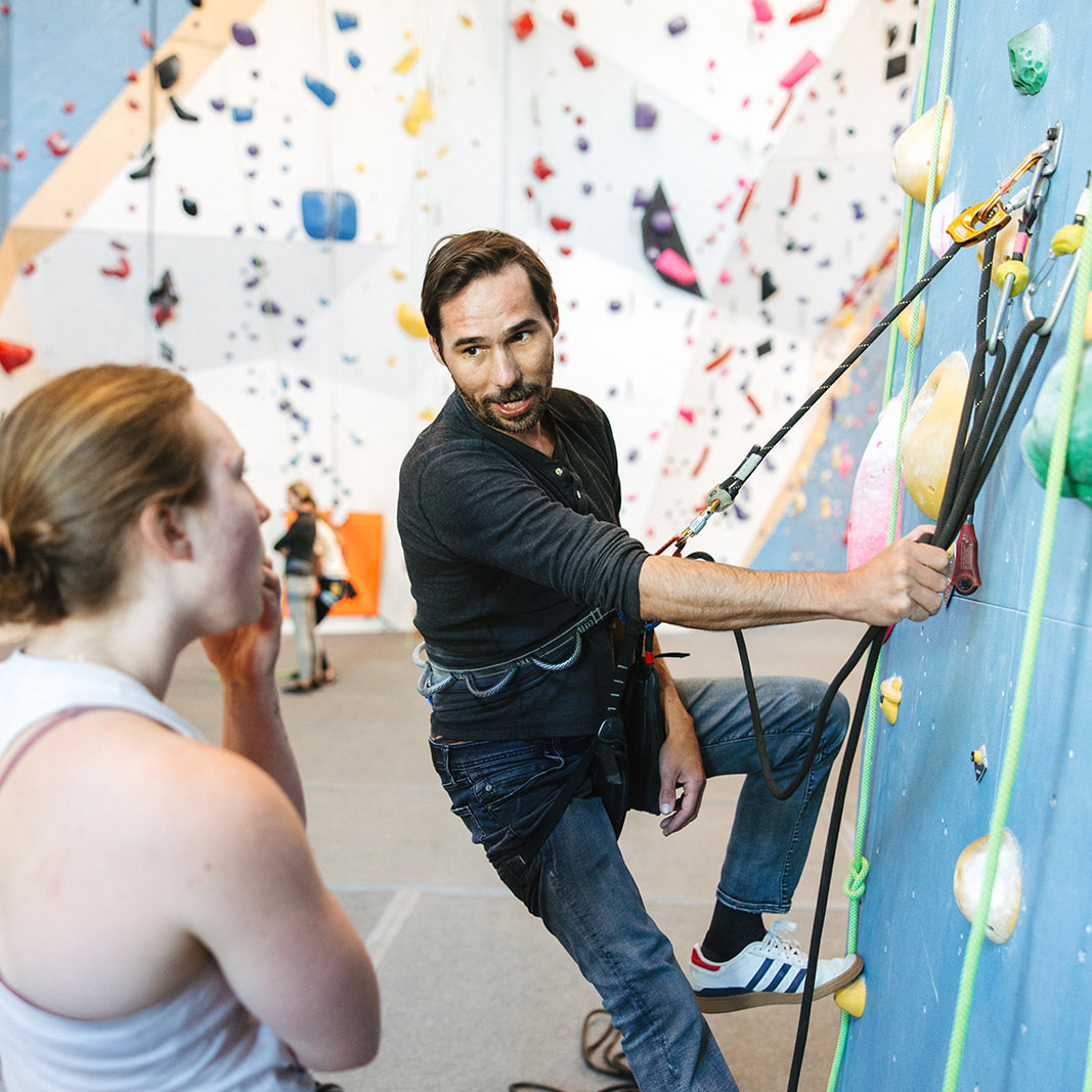 Session A man demonstrates a climbing technique on an indoor climbing wall while a woman in a harness watches and listens attentively. Bright climbing holds and ropes cover the wall in the background.
