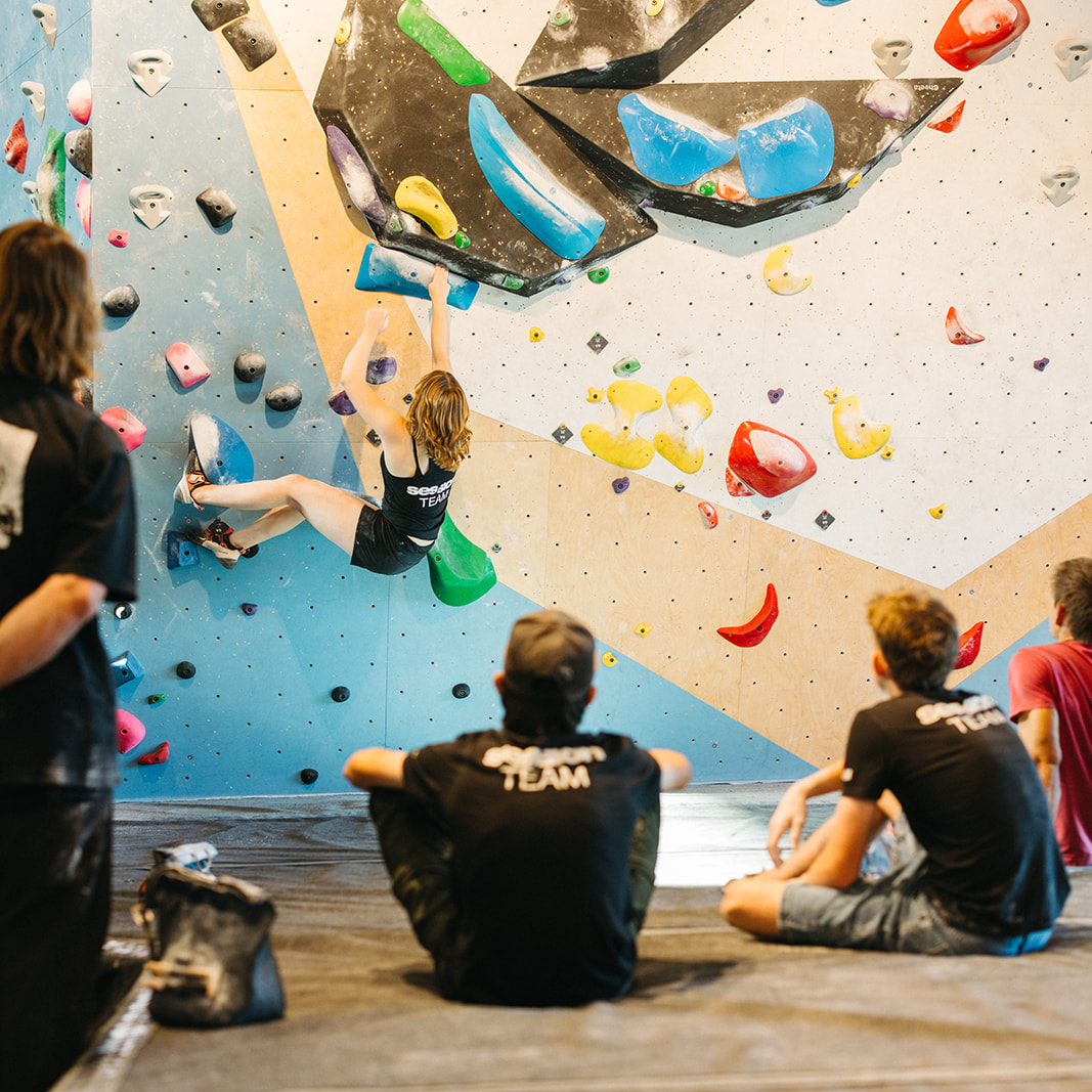 Session A youth team climber bouldering while teammates watch on in support.